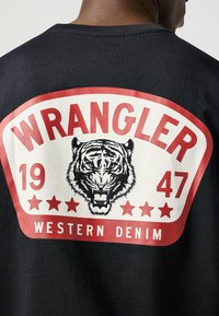 Camiseta negra con un gran gráfico en blanco y rojo. El diseño incluye un tigre, "WRANGLER" y "WESTERN DENIM" con estrellas.