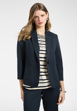 Kvinde med langt hår iført navyblazer og bukser, stribet top med udsmykninger, sølv hoop øreringe og armbånd mod en ensfarvet baggrund.