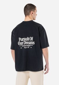 Fekete, oversized póló, hátul fehér szöveggel, amelyen a "Álmunk Után" felirat szerepel. Puha pamut anyagból készült, laza szabású.