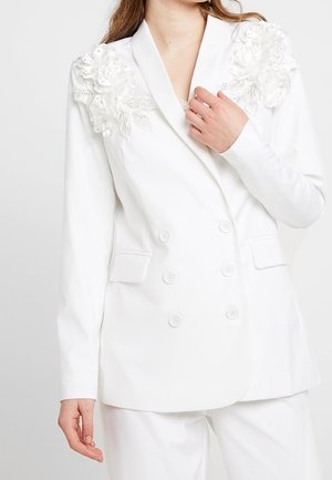 Blazer blanc à double boutonnage avec des appliqués floraux sur les épaules et quatre boutons à l'avant. Tissu lisse avec un design ajusté.