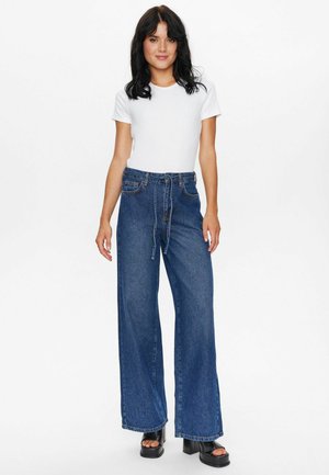 Weite Jeanshose mit hoher Taille, ausgestattet mit einem Kordelzug, in einer tiefblauen Waschung und mit gestickten Akzenten. Kombiniert mit einem weißen, taillierten T-Shirt.