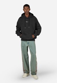 Čierna mikina s kapucňou a predným vreckom s bielym logom. Svetlo modré široké džínsy s bočnými pruhmi a zhrnutými manžetami. Biele tenisky.