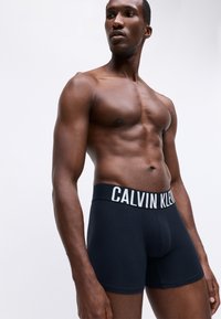 Calvin Klein fekete boxer alsók széles fehér logós derékkal. Puha pamut anyag, testhezálló kialakítás, comb középig érő hossz, minimalista stílus.