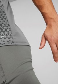 Maglietta sportiva grigia con un motivo geometricamente testurizzato sul lato. Il materiale è traspirante, etichettato "DRYCELL". Vista ravvicinata della mano e della vita.