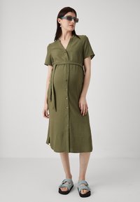 Olivenfarget midi-kjole med korte ermer, knappdetaljer og knyttet midje. Kombinert med blå sandaler med tykke stropper og plattformsåle.