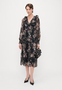 Čierne kvetinové šaty s dlhými priesvitnými rukávmi, volánikovým výstrihom a vrstveným dizajnom. Doplnene čiernou kabelkou a vysokými topánkami na opätku so zlatými akcentami.