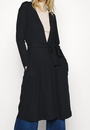 Kobieta w długim czarnym płaszczu z paskiem, nałożonym na beżową bluzkę i niebieskie dżinsy, ręce w kieszeniach płaszcza, stoi na białym tle.