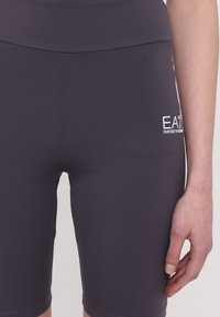 Szürke sportnadrág rugalmas anyagból, magas derékkal és egy kis fehér logóval az oldalon. Sima textúra, testhezálló fazon.