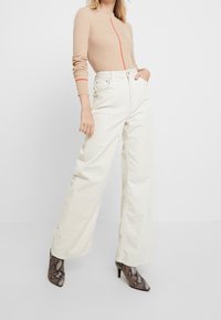 Haut beige à manches longues côtelé avec un détail de fermeture éclair rouge, associé à un pantalon large en velours côtelé blanc cassé taille haute et des talons noirs à imprimé serpent.