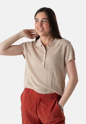 Joven mujer con cabello largo y oscuro que lleva una camisa beige de manga corta con cuello y pantalones rojos holgados, sonriendo y posando con una mano detrás de la cabeza.
