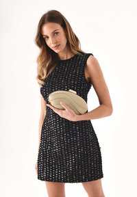 Abito nero con paillettes dalla silhouette aderente, completato da una pochette beige chiaro curva tenuta in mano. Texture liscia e design elegante.