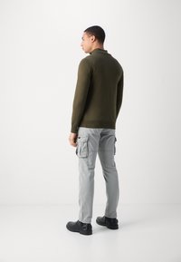 Polo à manches longues vert olive, associé à un pantalon cargo gris clair avec des poches latérales. Bottes noires avec une détail de boucle. Fond minimaliste.