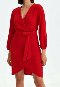 Robe rouge à manches longues, en tissu texturé, avec une ceinture à nouer à la taille. Présente un décolleté en V profond et un ourlet asymétrique.