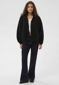 Sort fleecetrøje med lynlås foran, ribstrikkede manchetter og to sidelommer, kombineret med mørkeblå, udvidede jeans og sorte sko.