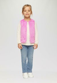 Gilet en fausse fourrure rose porté sur un haut à manches longues blanc, associé à un jean bleu et des baskets blanches. Enfant se tenant debout avec une expression neutre.