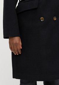 Dobbeltradet svart ullkåpe med to brune knapper, lommepatch og tilpasset passform, som viser en hånd som hviler på stoffet.
