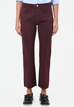 Donna che indossa pantaloni bordeaux cropped con orli grezzi e scarpe nere in pelle con lacci su sfondo bianco.
