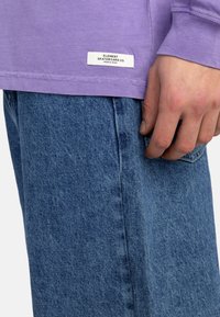 Lila Langarmshirt mit einem weißen Etikett, auf dem "ELEMENT SKATEBOARD CO." steht, kombiniert mit blauen Jeans im geraden Schnitt und feiner Textur.