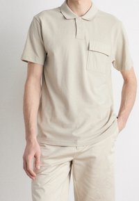 Polo shirt en coton beige avec un col, des manches courtes et une poche sur la poitrine. Le tissu a une texture lisse et une coupe décontractée.