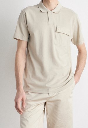 Polo shirt en coton beige avec un col, des manches courtes et une poche sur la poitrine. Le tissu a une texture lisse et une coupe décontractée.
