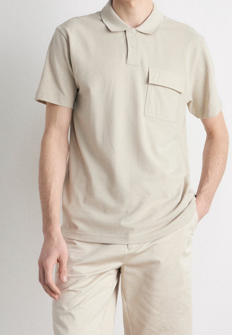 Polo shirt en coton beige avec un col, des manches courtes et une poche sur la poitrine. Le tissu a une texture lisse et une coupe décontractée.