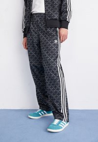Adidas träningsbyxor i svart med ett subtilt bladmönster, utrustade med vita sidostråk och resårmidja, tillsammans med gröna sneakers.