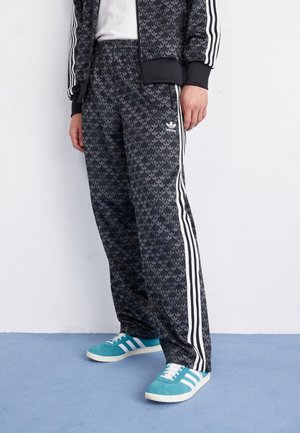 Zwarte Adidas trainingsbroek met een subtiele loofpatroon, voorzien van witte zijstrepen en een elastische tailleband, gecombineerd met teal sneakers.