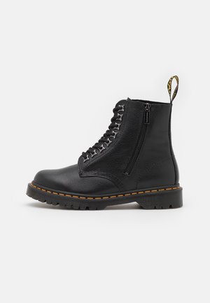 Bottine noire en cuir à lacets avec fermeture éclair sur le côté, semelle épaisse à crampons, surpiqûres jaunes et languette marquée à l'arrière.