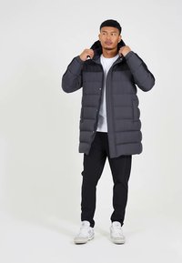 Tumši pelēks pūkains mētelis ar kapuci, izšūtu dizainu, spiedpogu aizdari, sānu kabatām, uzvilkts pār baltu krekls un melnām biksēm, kopā ar baltām sporta kurpēm.