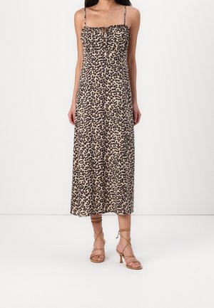 Frau trägt ein ärmelloses Midikleid mit Leopardenmuster, dünnen Trägern und beigefarbene, geschnürte Sandaletten mit Absatz vor weißem Hintergrund.