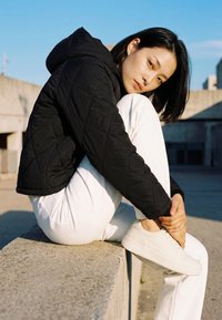 Jonge vrouw in een zwart doorgestikt jack en witte broek zit op een betonnen rand, houdend haar enkel vast buiten met een blauwe lucht op de achtergrond.