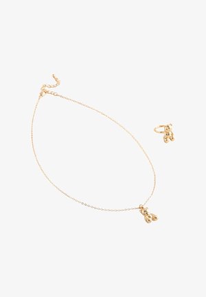 Goldenes Halsketten- und Ring-Set, jeweils mit einem kleinen polierten Teddy-Bär-Anhänger an einer feinen Kette und einem Band vor einem weißen Hintergrund.