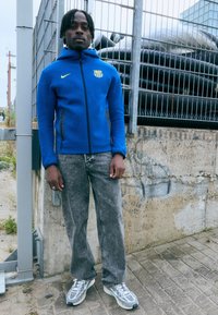 Μπλέ φούτερ με κουκούλα και κίτρινο λογότυπο, γκρι τζιν παντελόνι και γκρι αθλητικά παπούτσια με μπλέ λεπτομέρειες. Το σκηνικό περιλαμβάνει έναν τσιμεντένιο τοίχο και έναν φράκτη.