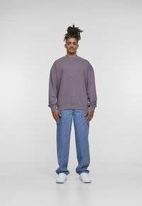 Felpa viola con collo a giro e spalle ribassate, abbinata a jeans dritti blu chiaro e sneakers bianche. Sfondo semplice.