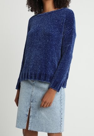 Kobieta ubrana w niebieski sweter z długim rękawem o teksturowanej powierzchni oraz jasnoniebieską spódnicę dżinsową z przednim rozcięciem, stojąca na jednolitym tle.