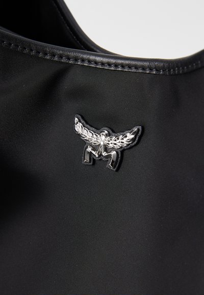 Emblema metálico prateado com design de asas e aranha em tecido preto texturizado com borda de couro costurada.