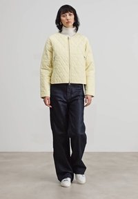 Gep quiltete gelbe Jacke mit Reißverschluss, cropped Design, langen Ärmeln und zwei seitlichen Taschen, getragen über einem weißen Rollkragenpullover und kombiniert mit dunklen Jeans.