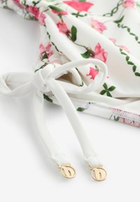 Blumendekorierter Stoff mit rosa Rosen und grünen Blättern auf weißem Hintergrund, mit weißen Trägern und goldenen runden Hardware-Akzenten.