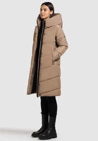 Kvinde iført en lang beige pufferjakke med høj krave, sorte leggings og sorte chunky støvler, stående på siden foran en lys baggrund.