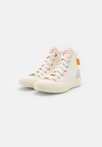 Zapatillas de lona de caña alta, color blanco roto, con cordones en la parte delantera, suelas de goma y etiqueta naranja en el talón. Presenta una marca roja en la lengüeta.