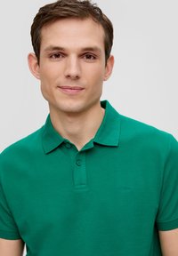 Zielona koszulka polo wykonana z teksturowanej tkaniny, z klasycznym kołnierzykiem, zapięciem na trzy guziki oraz haftowanym logo na piersi.