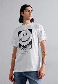Hvit bomull t-skjorte med et stort svart smilefjesgrafikk og "MOSCHINO" tekst, sammen med lyseblå denimjeans.