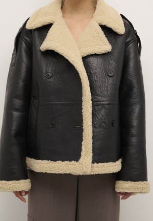 Sort læderjakke med cremefarvet shearling-krave, manchetter og kant, dobbeltspændt med fire sorte knapper, båret over brune bukser.