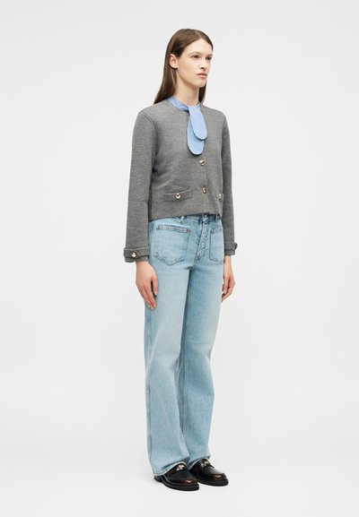 Γυναίκα που στέκεται φορώντας γκρι σακάκι με κουμπιά, ριγέ μπλε γραβάτα, ανοιχτό μπλε τζιν και μαύρα μοκασίνια σε απλό λευκό φόντο.