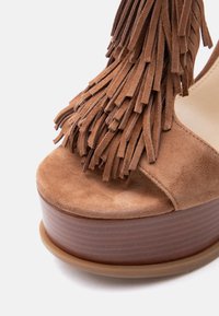 Καφέ πλατφόρμα σανδάλι από σαουέτ με στρωματωτά φουντάκια πάνω από το ανοιχτό δάχτυλο και σόλα από στοιβαγμένο ξύλο.