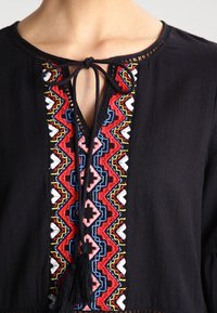 Top nero con vivaci ricami multicolori lungo il colletto, motivi geometrici e chiusura a cravatta con un accento a nappina.