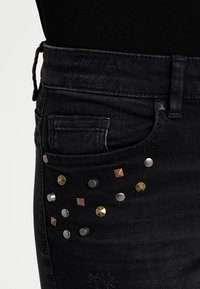 Svarta denimjeans med en närbild av en ficka dekorerad med olika metallspikar i guld-, silver- och bronsfinish.