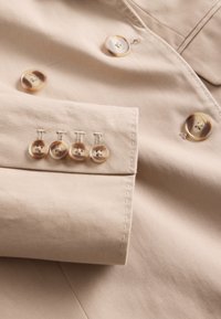 Bež tkanina s prikazom krojenog dizajna s pet kontrastnih gumba na manžeti i dodatnim gumbima sprijeda. Uređen šav s detaljima.