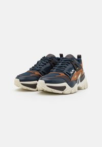Zapatillas gruesas en azul marino y marrón con cordones negros y suelas blancas gruesas, colocadas una al lado de la otra sobre un fondo blanco.