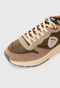 Sneaker din suede maro și bej cu sireturi crem, patch cu brand pe lateral și talpă bej pe un fundal alb.